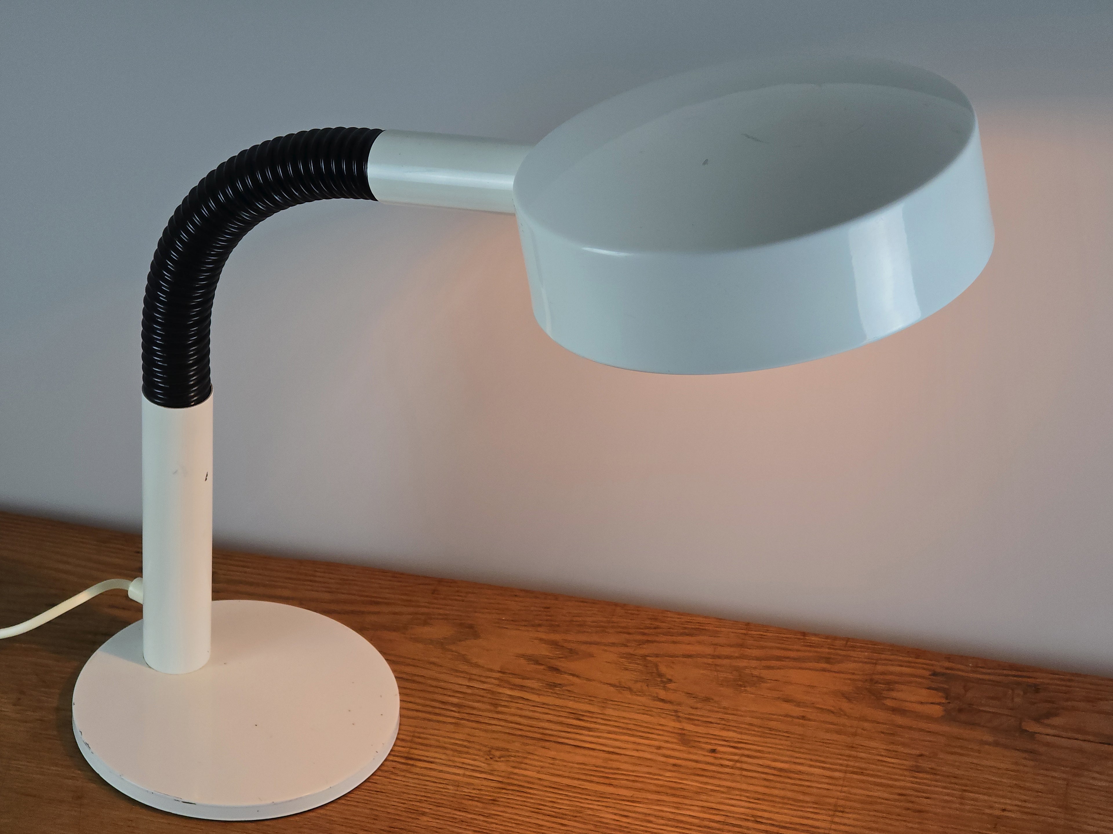 1960s Mid Century Modern gooseneck desk lamp by Hala Zeist, bird's eye view from the top side, showing the design of the round white metal shade, the lamp is lit, on a vintage wooden table.