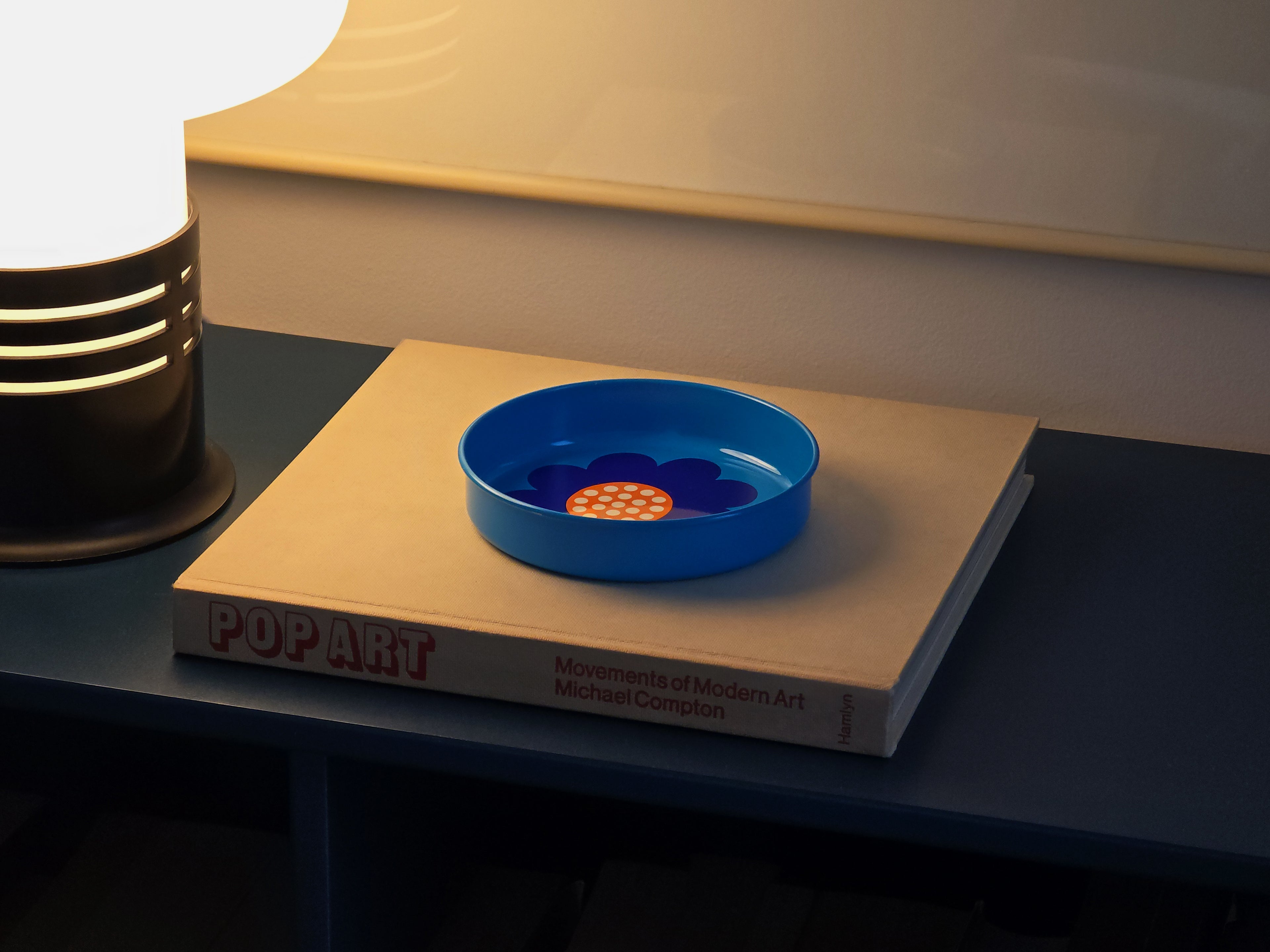 Perspective image of a vintage 1970s Laurids Lønborg tin tray designed by Al and Lena Eklund, styled image on a book, set on a blue cabinet, with a mushroom lamp lit next to it.