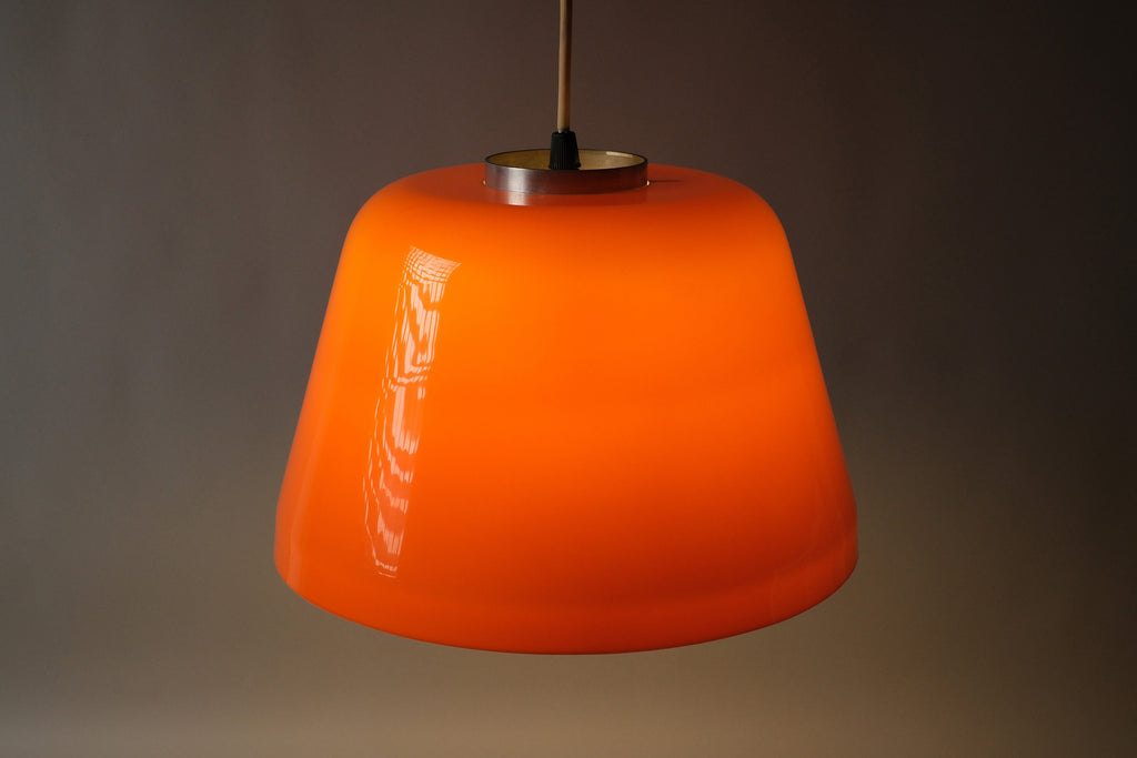 Front illuminated view of 1970s dual-layer orange acrylic Space Age pendant lamp, showing warm diffused downward light through the white interior layer against a dark background.