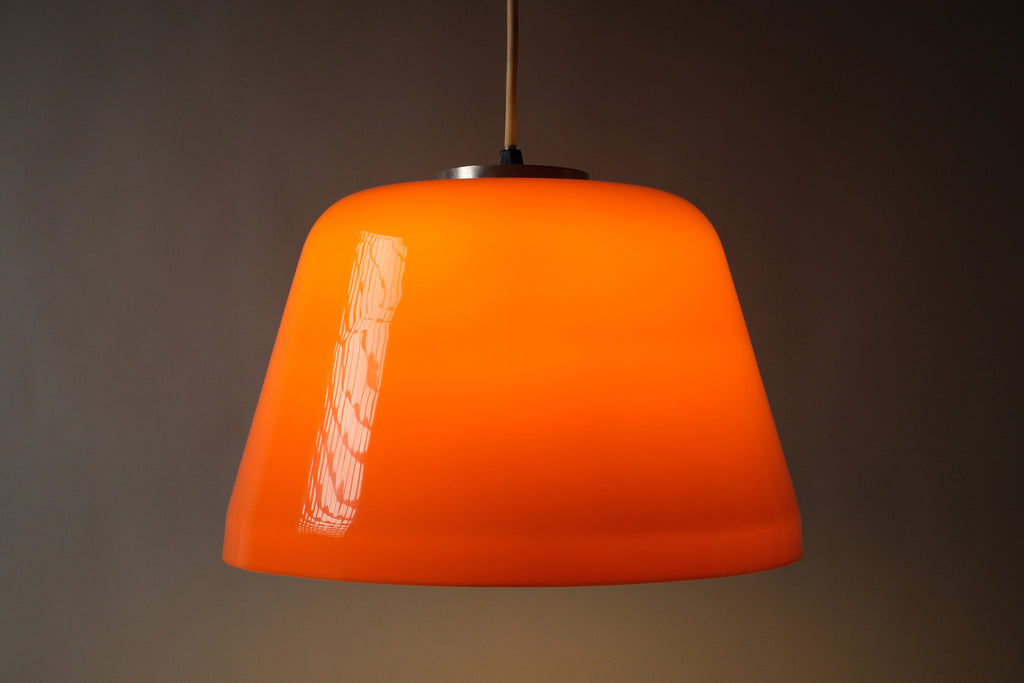 Full profile illuminated view of a 1970s dual-layer orange acrylic Space Age pendant lamp, showing the warm glow emanating from the white interior layer with the cord and ceiling cap visible.