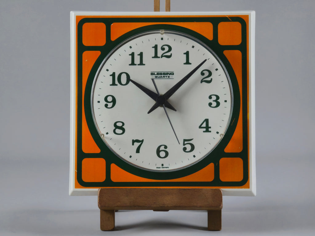 1970s Space-Age Design Blessing orange plastic wall clock, front view with the clock on a wooden stand and a neutral light grey background.