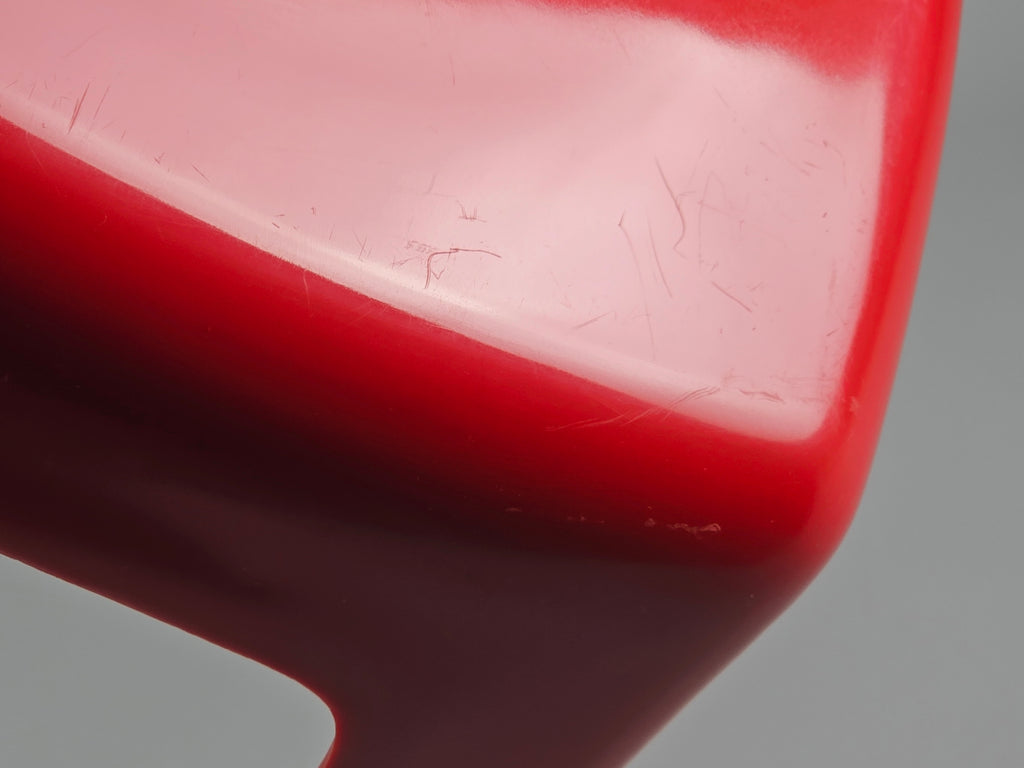May include: Close-up of a glossy red object with a curved shape. The surface has minor scratches and imperfections. The object's colour transitions from a lighter red to a darker shade, creating a gradient effect. The background is a neutral grey.