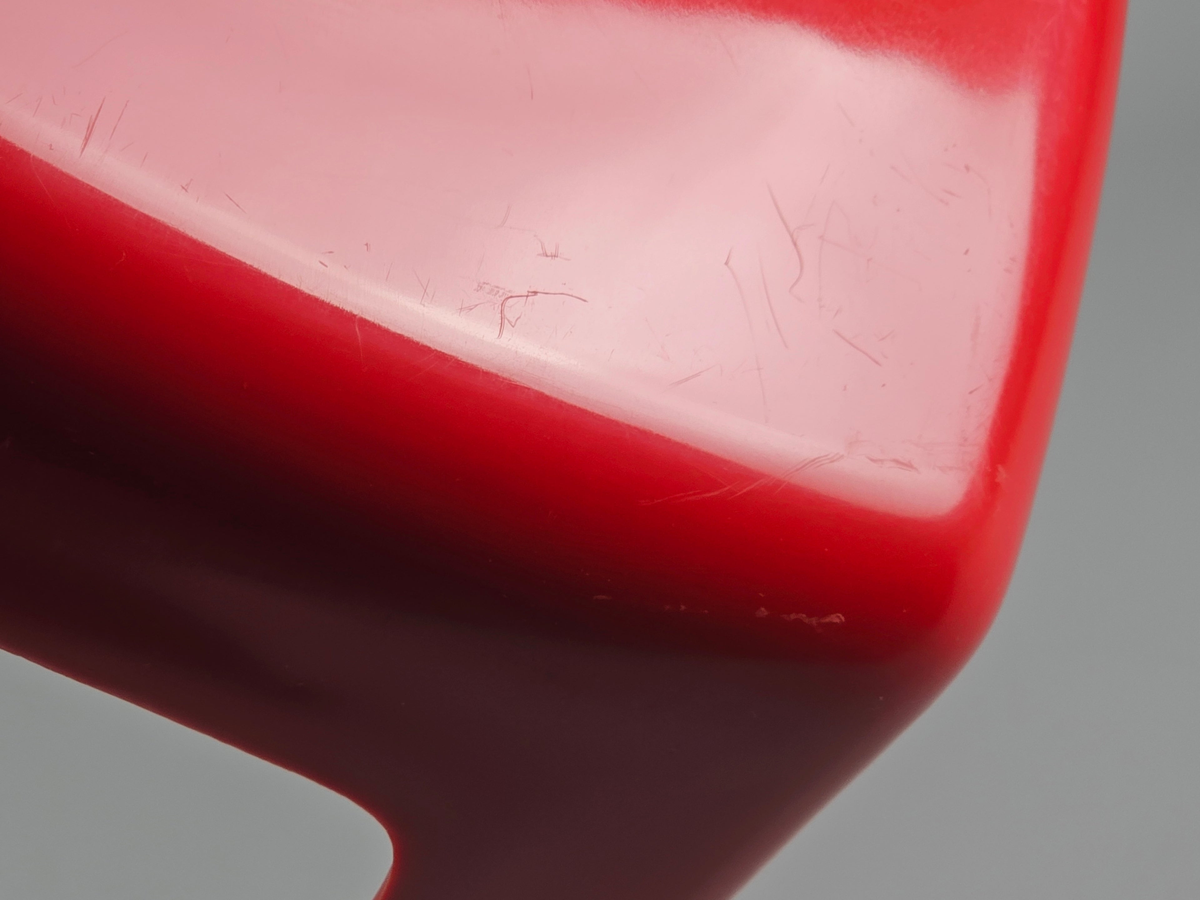 May include: Close-up of a glossy red object with a curved shape. The surface has minor scratches and imperfections. The object's colour transitions from a lighter red to a darker shade, creating a gradient effect. The background is a neutral grey.
