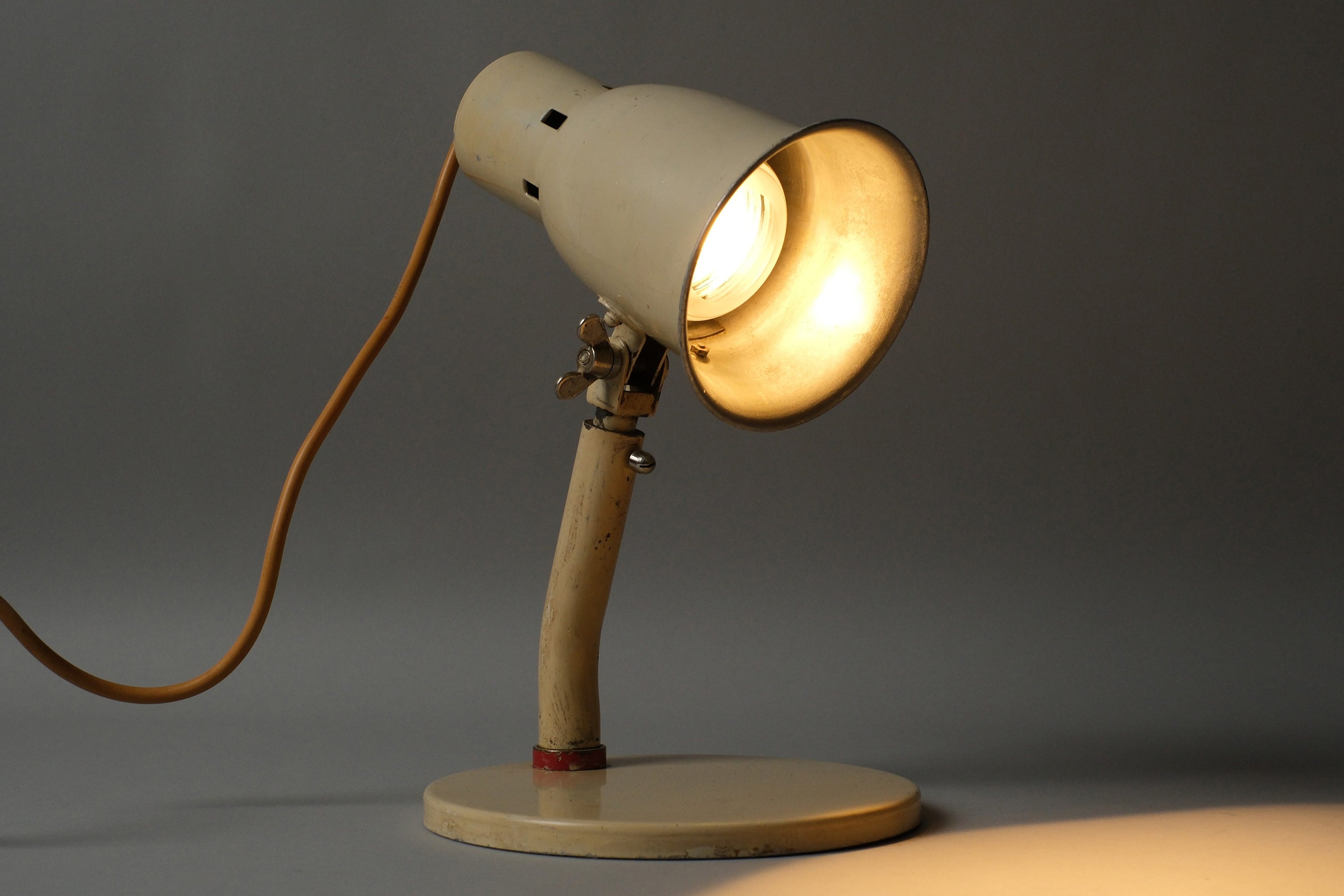 Front illuminated view of a 1920s-1930s Bauhaus industrial desk lamp in cream enamel, Kandem type, casting warm directional light from the adjustable shade.