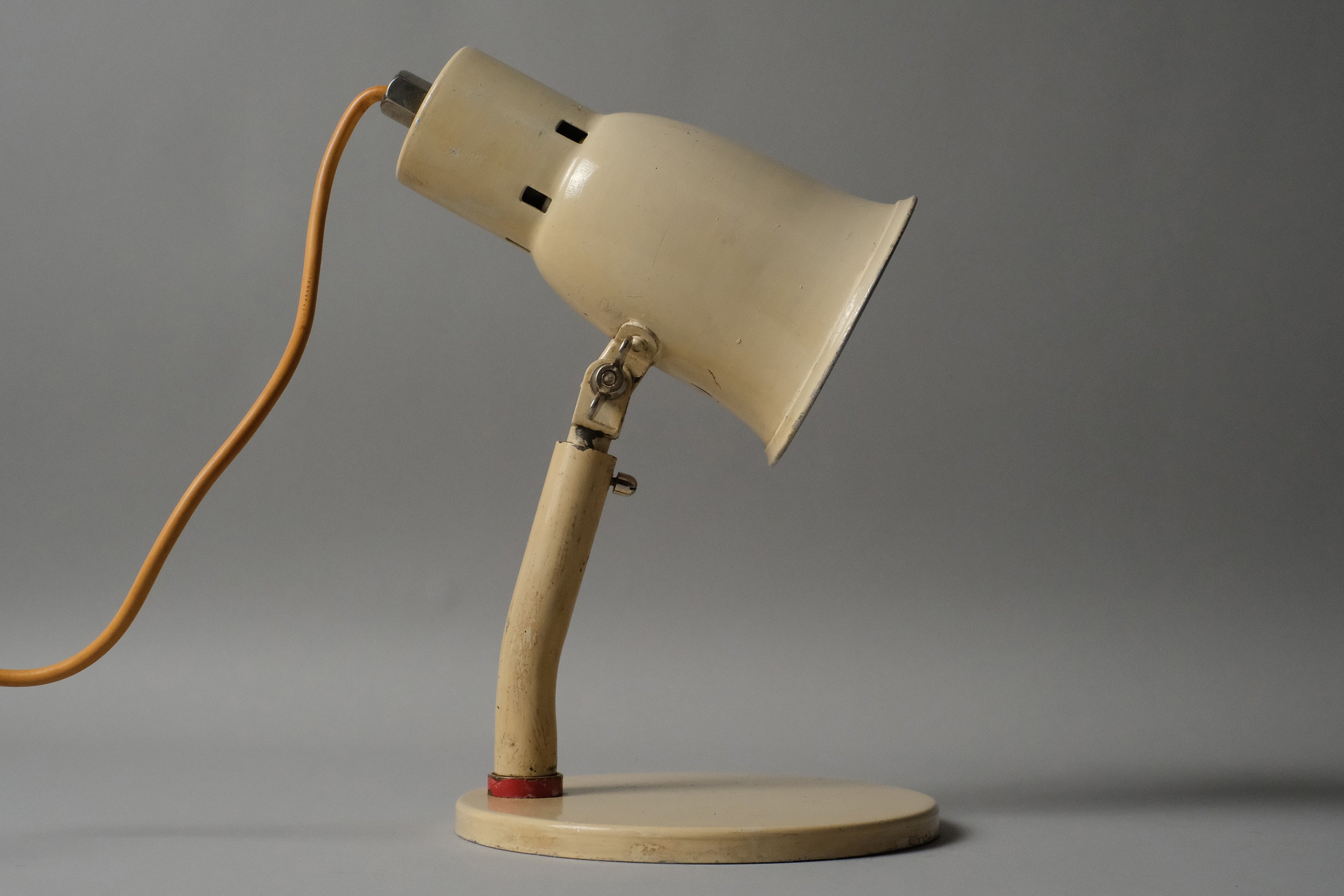 Profile view of a 1920s-1930s Bauhaus industrial desk lamp in cream enamel, Kandem type, showing the double pivot articulation and cast iron oval base against a dark background.