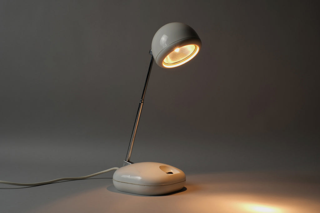 Illuminated view of a 1970s Hustadt Leuchten antenna halogen desk lamp from West Germany, showing warm directional halogen light cast from the spherical spot head on the extended antenna stem.