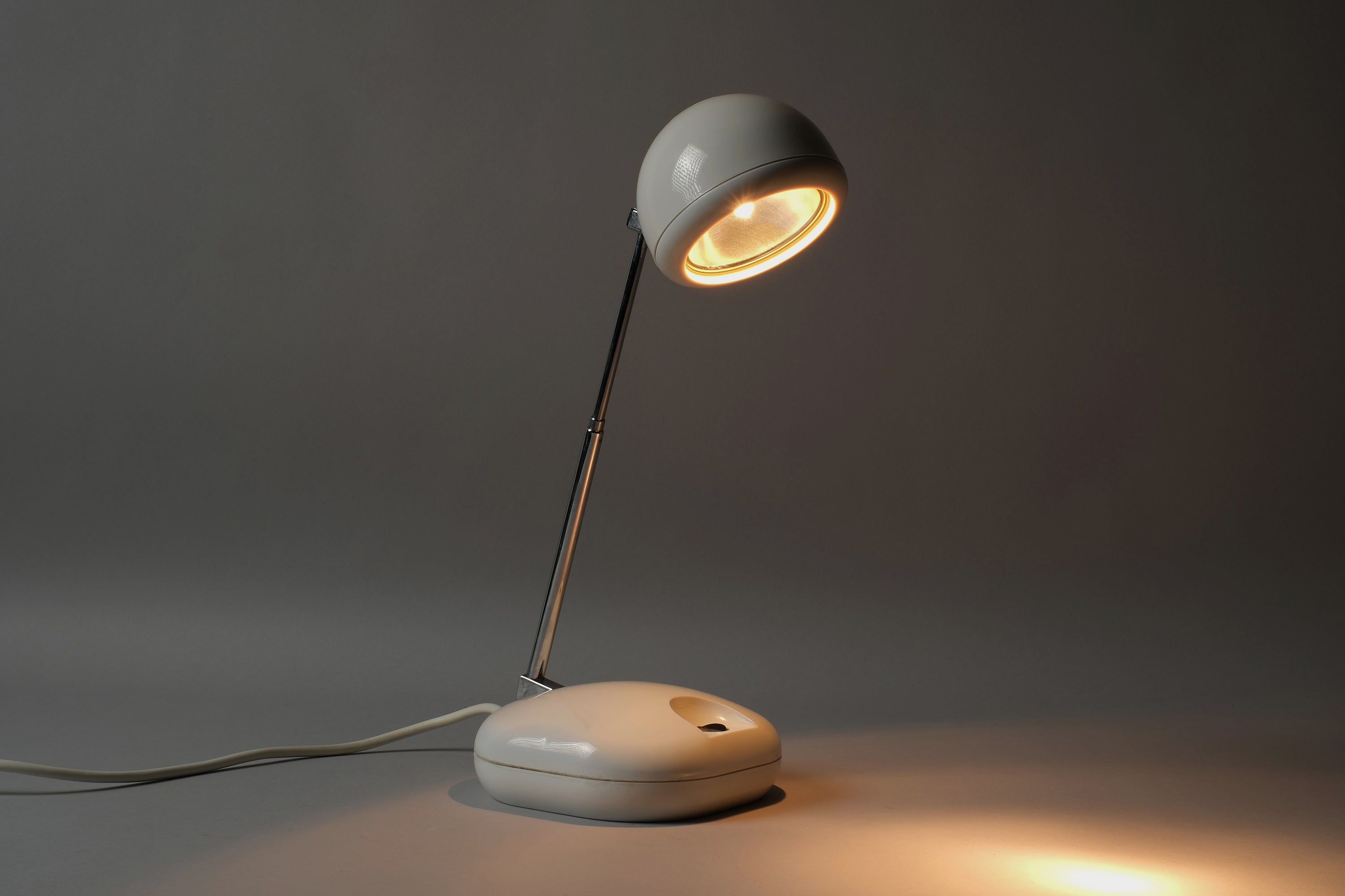Illuminated view of a 1970s Hustadt Leuchten antenna halogen desk lamp from West Germany, showing warm directional halogen light cast from the spherical spot head on the extended antenna stem.