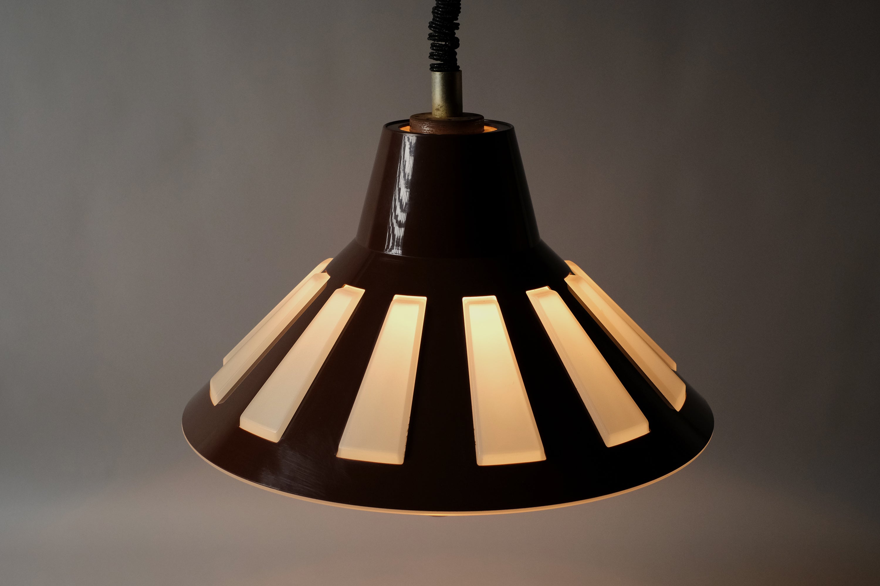 Angled illuminated view of a 1970s Massive Lighting Belgium UFO pendant lamp, showing the geometric cutout pattern in the brown outer shell casting warm striped light through the translucent inner layer.