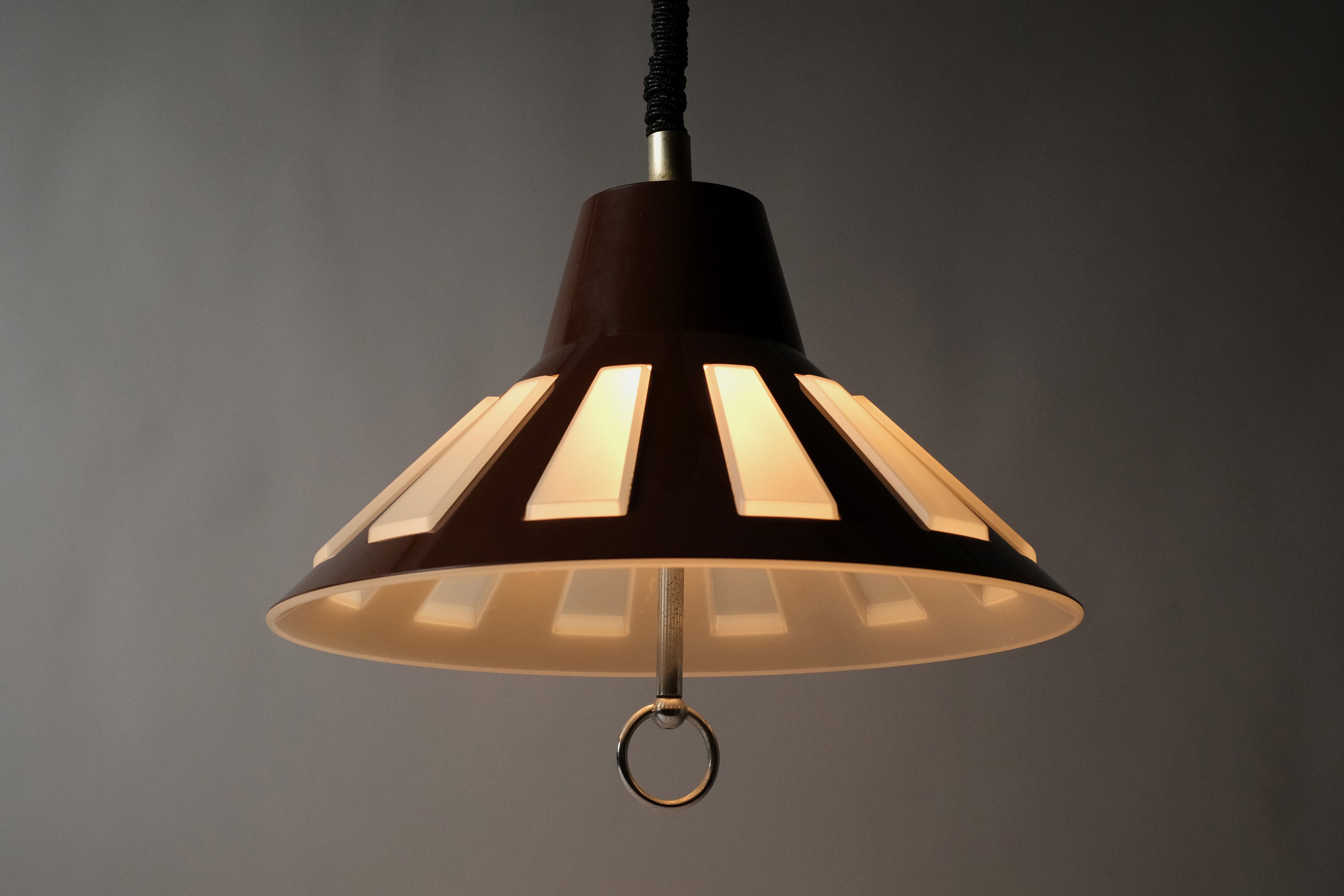 Front illuminated view of a 1970s Massive Lighting Belgium UFO pendant lamp, showing the warm striped light pattern produced by the brown outer ABS shell with geometric cutouts over the translucent inner diffuser.