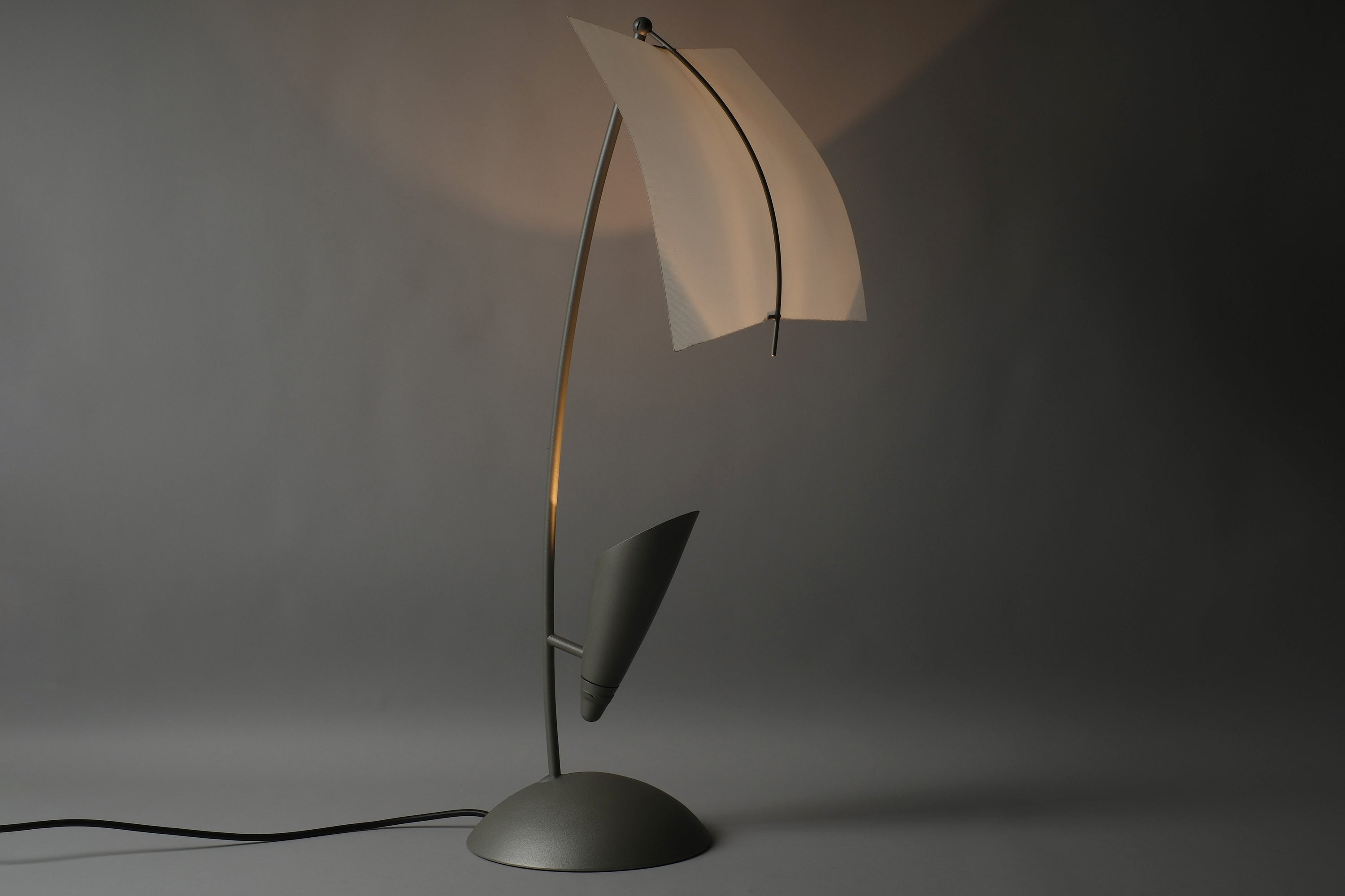 Rolf Benz Nuvola table lamp, Art. Nr. 79101009, illuminated in darkness, showing the curved white lacquered steel leaf diffuser glowing warm amber against a pure black background, Germany.
