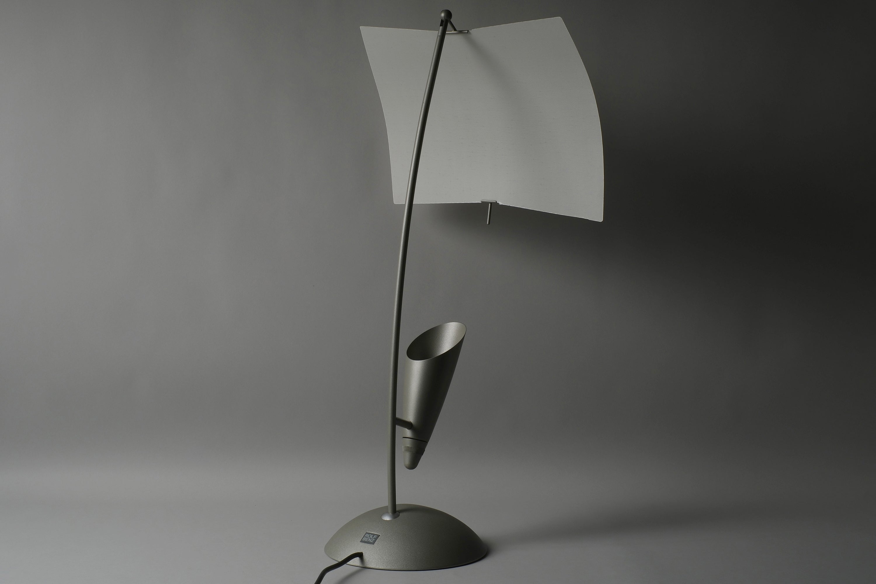 Full view of a Rolf Benz Nuvola table lamp, Art. Nr. 79101009, showing the curved white lacquered steel leaf diffuser on the arched stem with counterweight fin and matte grey dome base, Germany.