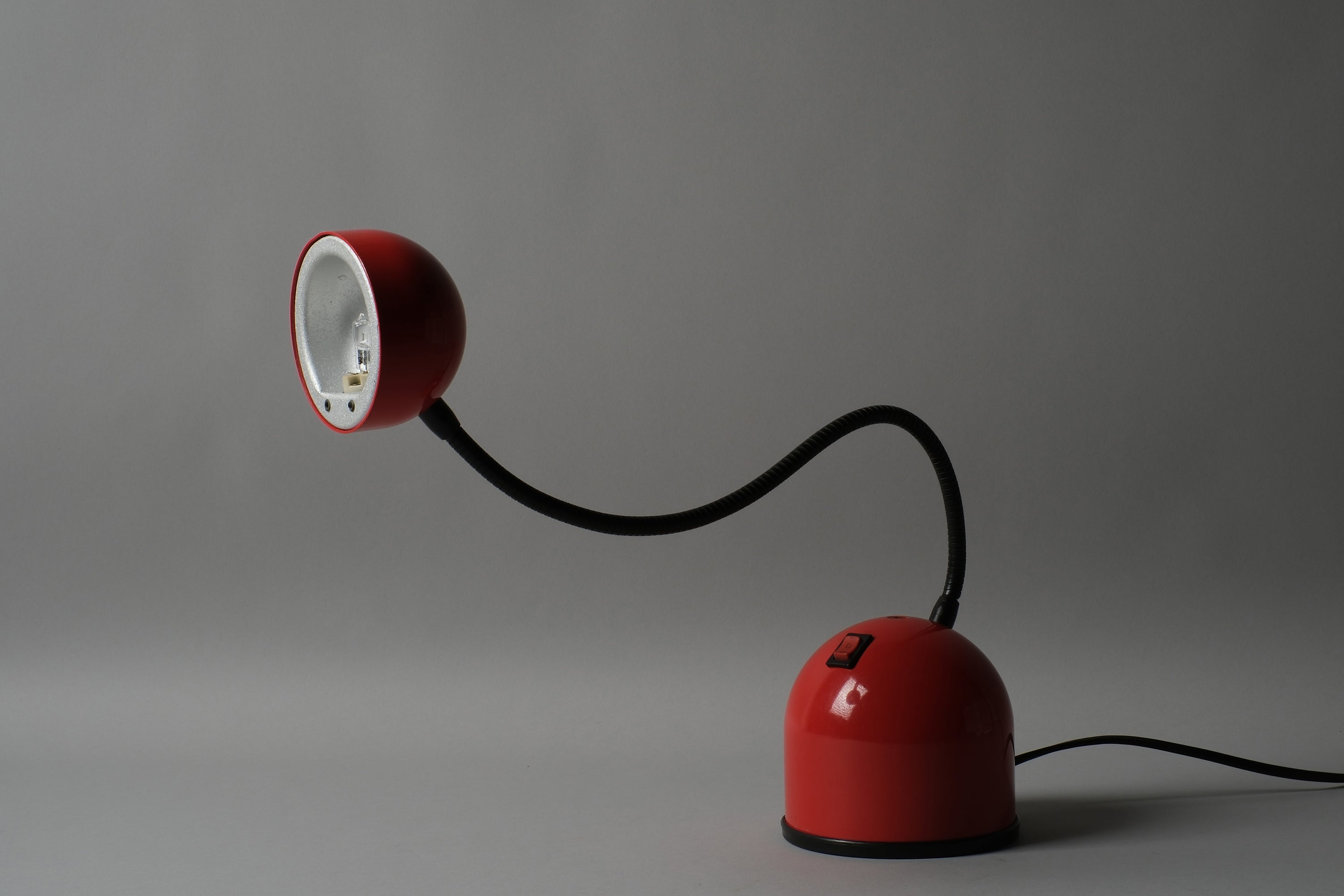 Perspective view of a 1980s Stilplast flexible halogen desk lamp from Italy, showing the red ABS dome base, black flexible gooseneck arm and small halogen spot head against a neutral background.