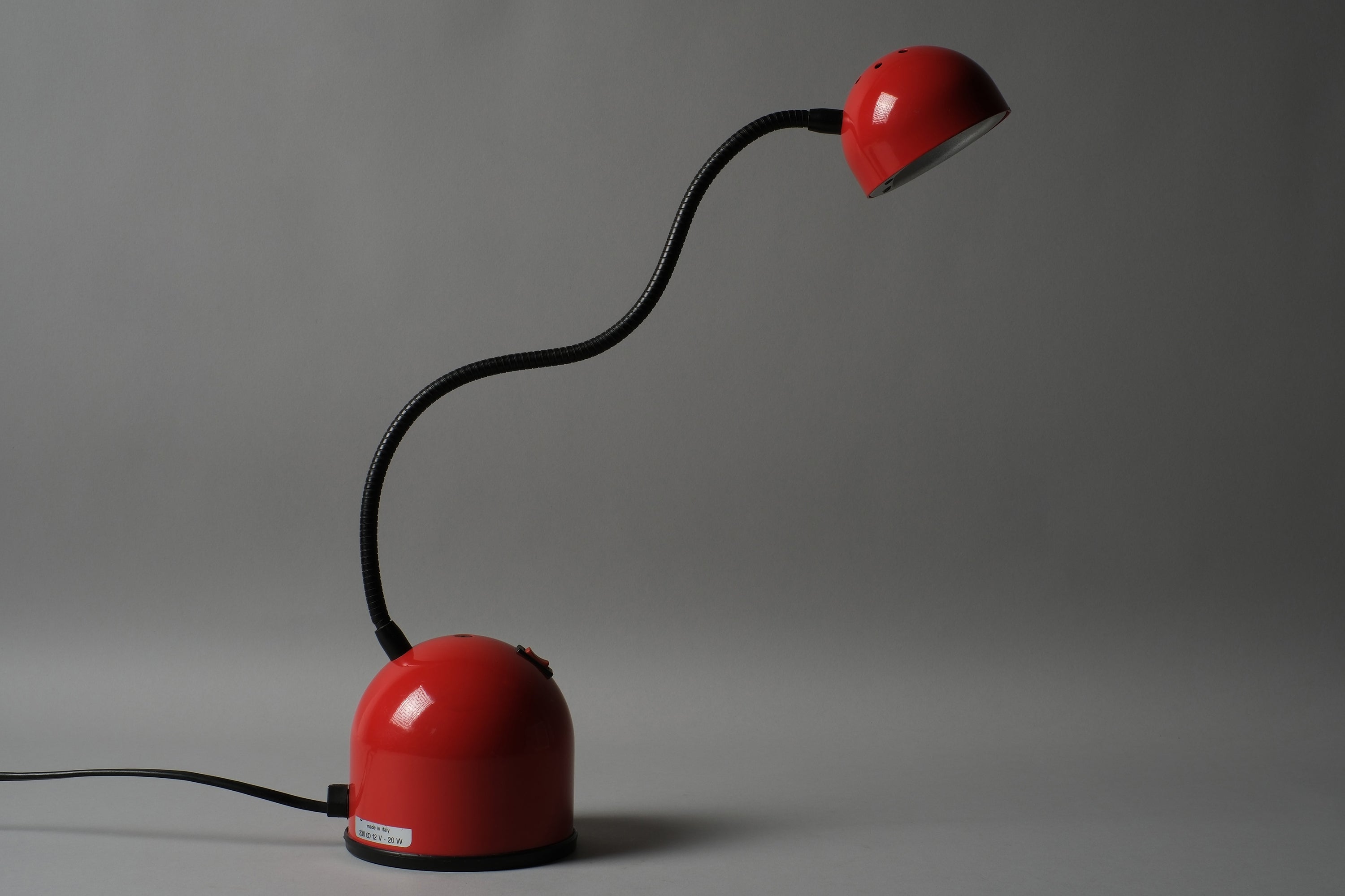 Full view of a 1980s Stilplast flexible halogen desk lamp from Italy in red ABS with black gooseneck arm, shown in profile against a dark background highlighting the Space Age form.