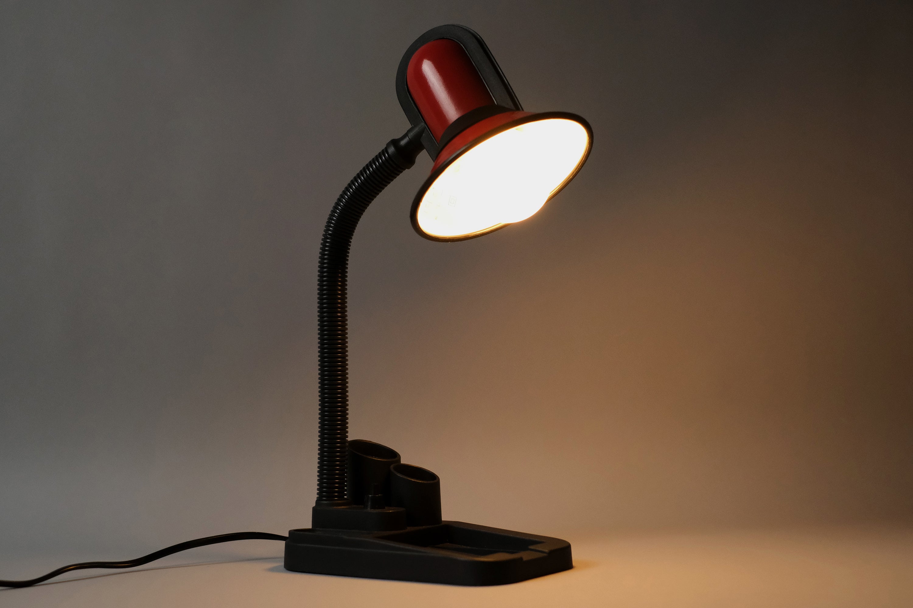 Angled and amber-lit view of the Fase gooseneck desk lamp from the 1980s, laid on a table with a light grey neutral background.