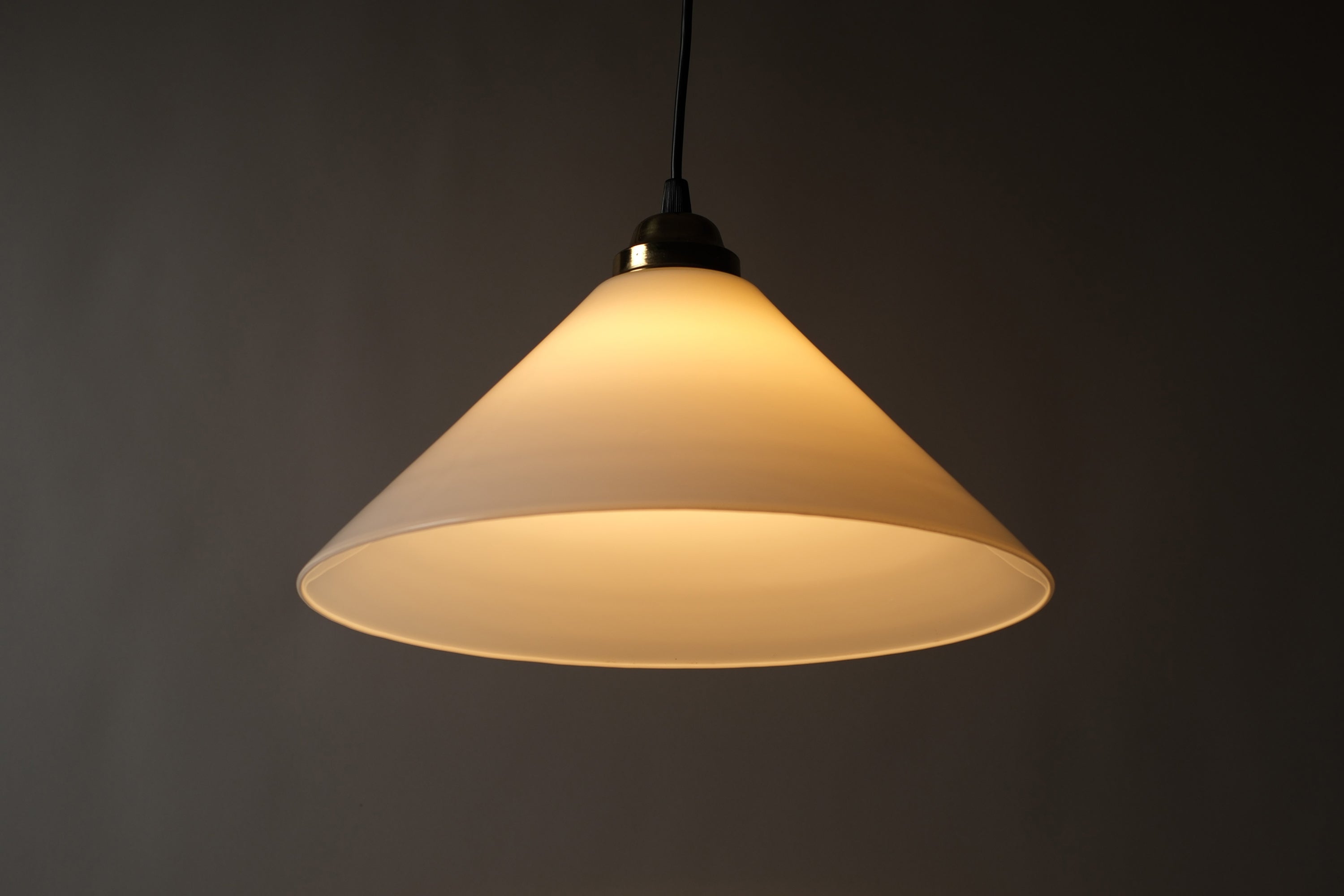 Front illuminated view of a 1980s vintage opal glass cone pendant lamp, showing the warm downward and outward glow cast through the opaline glass cone against a dark background.