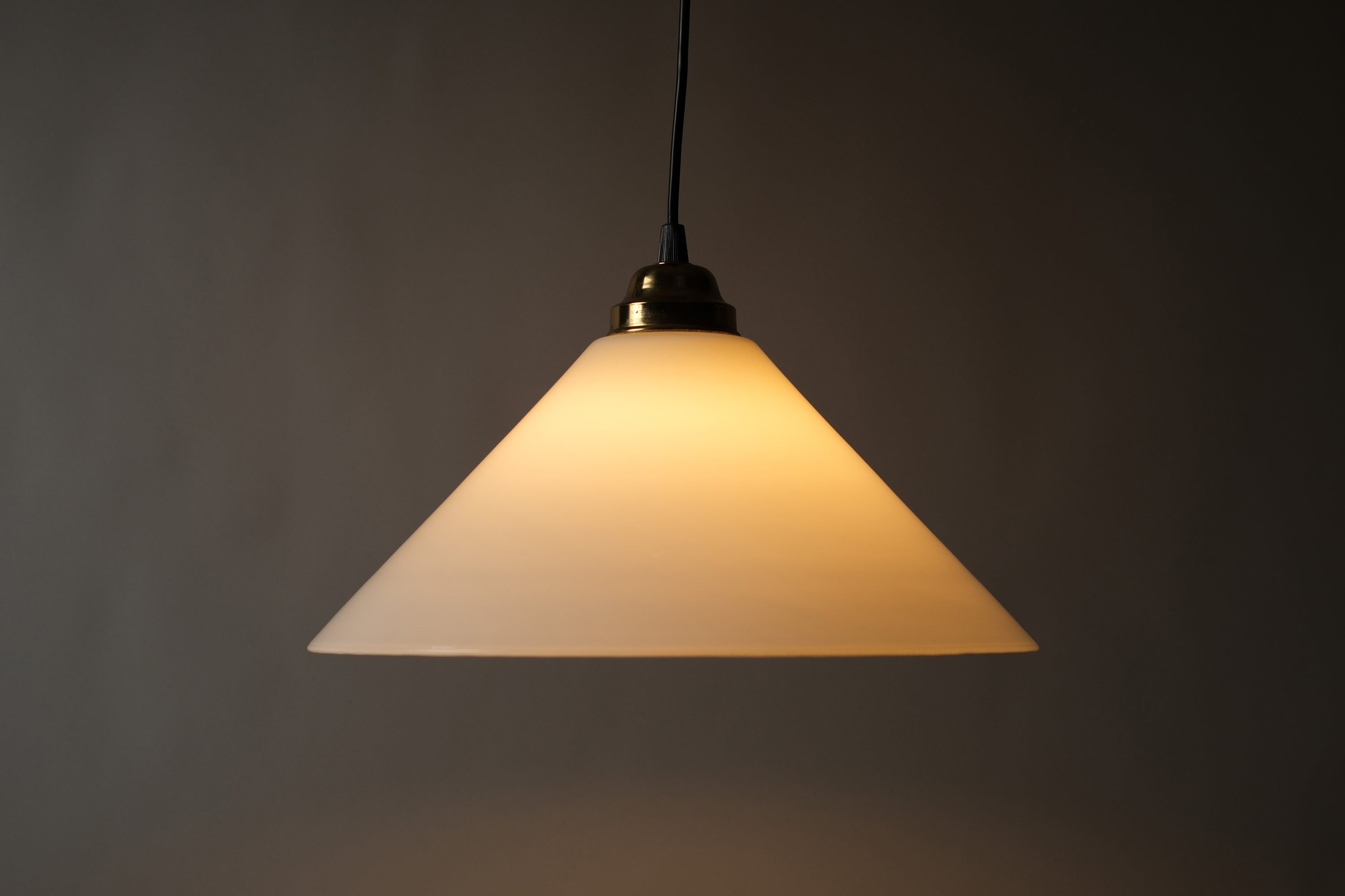 Profile illuminated view of a 1980s vintage opal glass cone pendant lamp, showing the warm amber glow radiating through the opaline glass cone and the clean conical silhouette when lit.