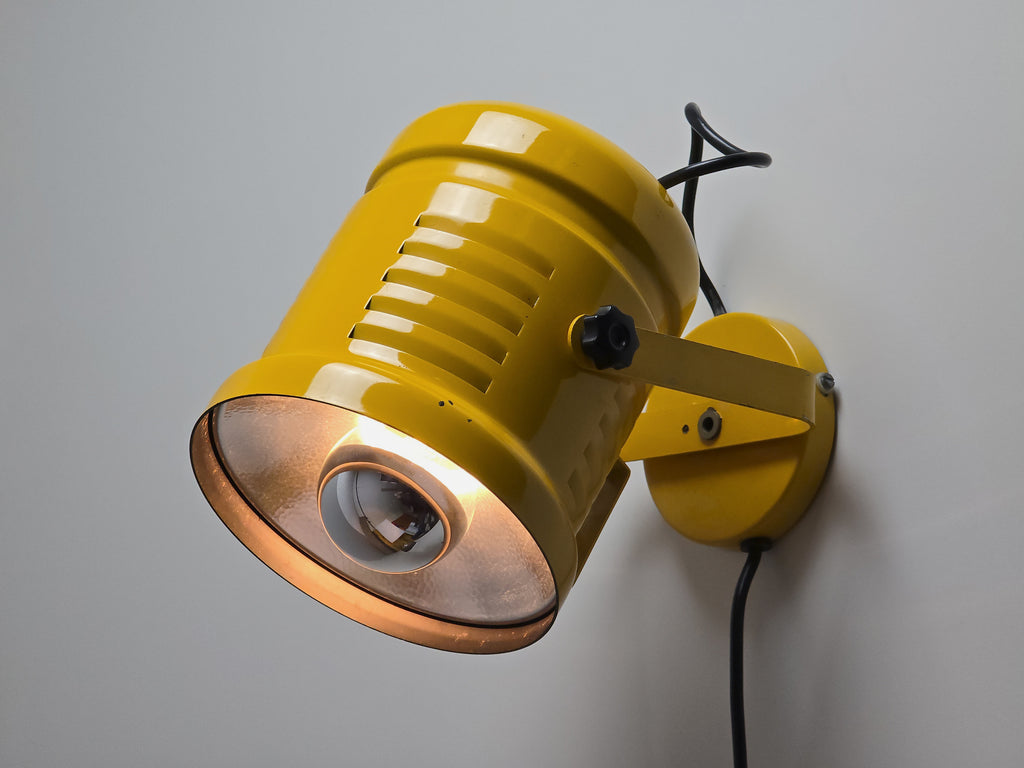 Amber lit view of vintage 1980s industrial wall lamp in yellow enameled metal with adjustable knobs, wall mounted on a neutral background with black cord and a mirror bulb installed on it.