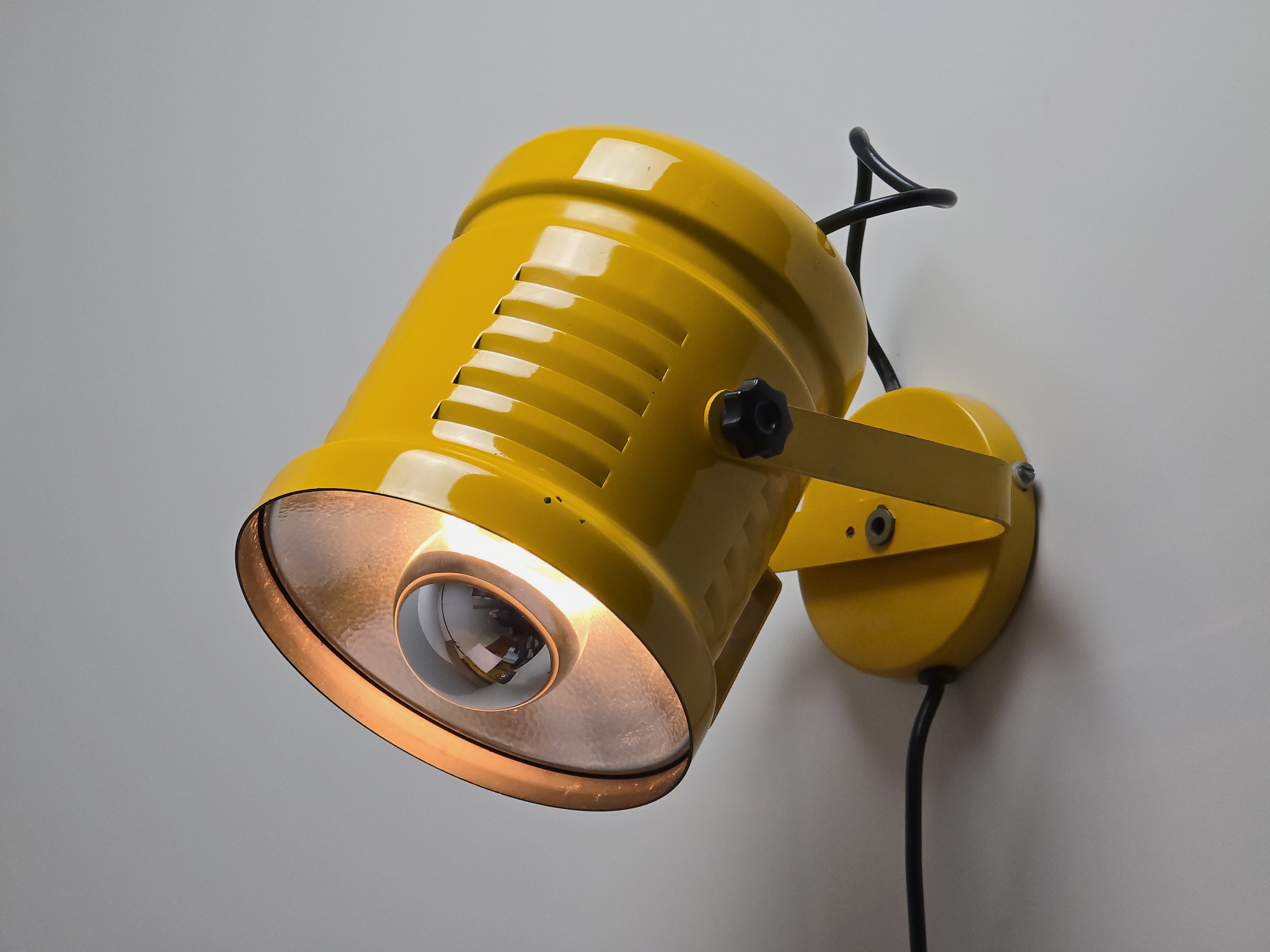 Amber lit view of vintage 1980s industrial wall lamp in yellow enameled metal with adjustable knobs, wall mounted on a neutral background with black cord and a mirror bulb installed on it.